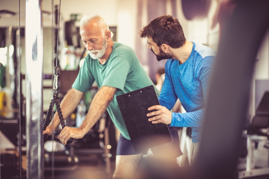 Fit im Alter: Mann trainiert seine Arme im Fitnessstudio unter Aufsicht eines Trainers